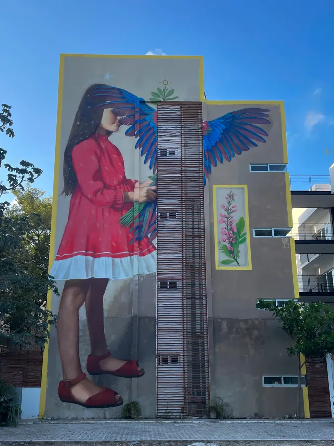 Mural de Bache: niña con vestido rojo y alas de guacamaya en la fachada de un edificio en Tulum