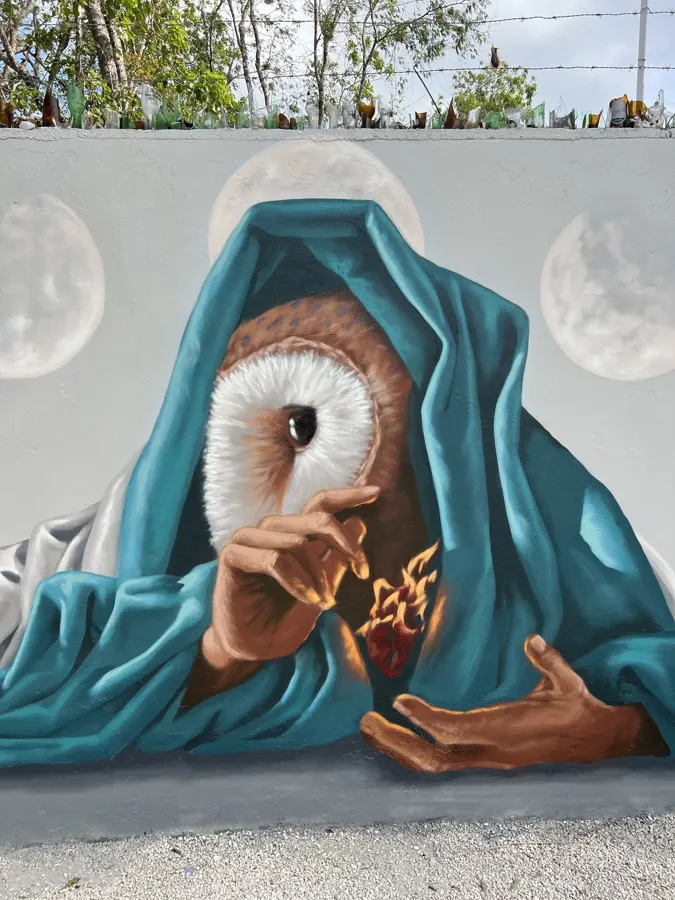 Mural de Secreto: figura con rostro de búho y manto turquesa sosteniendo un corazón en llamas